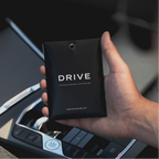 Hand holding a black 'Drive' car perfume packaging inside a vehicle.